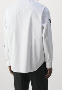 Weißes Langarmshirt aus Baumwolle mit abgerundetem Saum, geknöpften Manschetten und glatter Textur. Die Rückansicht zeigt minimale Details.