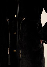 Abrigo de piel sintética negra con botones dorados y cordones negros con borlas. El abrigo tiene una textura suave y una silueta relajada.