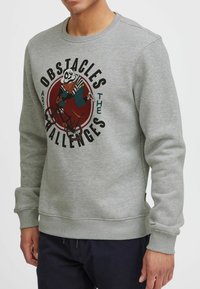 Vaaleanharmaa collegepaita, joka on valmistettu pehmeästä kankaasta ja jossa on värikäs kuva pyörällä ajavasta hahmosta sekä ympärillä rohkeaa tekstiä.