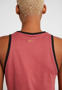 Personne aux cheveux blonds courts et bouclés portant un haut rouge sans manches avec des bordures noires et un petit logo Nike doré dans le haut du dos.