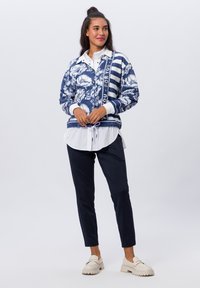 Navy blazer met bloemenpatroon en gestreepte accenten, daaronder een witte overhemd, gecombineerd met navy broek en crèmekleurige klompen.