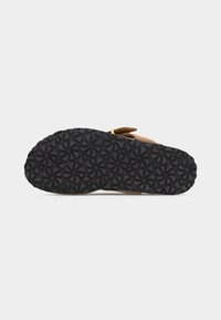 Semelle en caoutchouc noir avec motif de bande de roulement géométrique circulaire sur sandale à bride beige, vue de dessous sur fond clair.