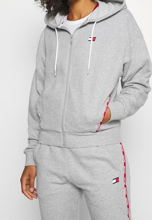 Person trägt einen hellgrauen Kapuzenpullover mit Reißverschluss und passende Jogginghose mit roten und weißen Markenzeichen-Details an den Taschen und am Hosenbein.