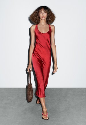 Red satin dress with a scoop neckline, fitted design, and ankle length. Large brown beaded bag and pink flip-flops complete the look.