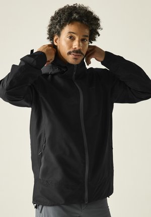 Man with curly hair adjusting the hood of a zipped black jacket, standing against a plain light background.