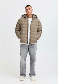En pufferjakke i dæmpet beige med lodret quiltet tekstur, lynlås og justerbar hætte. Parret med falmede grå jeans og hvide sneakers.