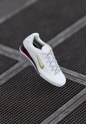 Sapato desportivo branco com parte superior texturizada, logo swoosh dourado, detalhes em vermelho no calcanhar e atacadores cinza. Sola elevada para amortecimento.