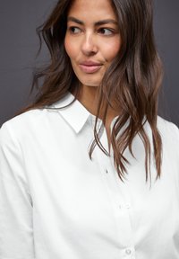 Chemise blanche à boutons avec un col classique et des manches longues. Le tissu semble lisse, avec une coupe décontractée et un subtil détail de bouton.