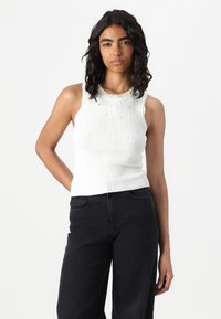 Nuori nainen, jolla on laineikkaat tummat hiukset, pukeutuneena hihattomaan valkoiseen ribbipaitaan, jossa on helmikoristeita, ja mustiin korkeavyötäröisiin housuihin valkoista taustaa vasten.