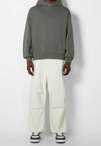Persona che indossa un ampio hoodie grigio, pantaloni bianchi larghi e sneakers bianco e nere, in piedi contro uno sfondo bianco semplice.