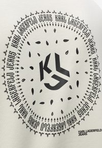 Bela tkanina z okroglo črno grafiko, ki vključuje logotip "KL" in dekorativne elemente, vključno s kapljastimi motivi in tekstualnimi poudarki.