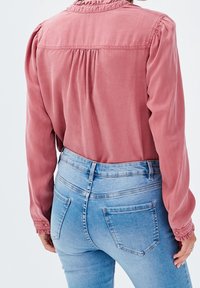 Langärmlige Bluse in Blush-Pink mit gerafften Schultern und Ausschnitt, kombiniert mit hoch sitzenden, hellblauen Jeans, die über Gesäßtaschen und eine glatte Oberfläche verfügen.