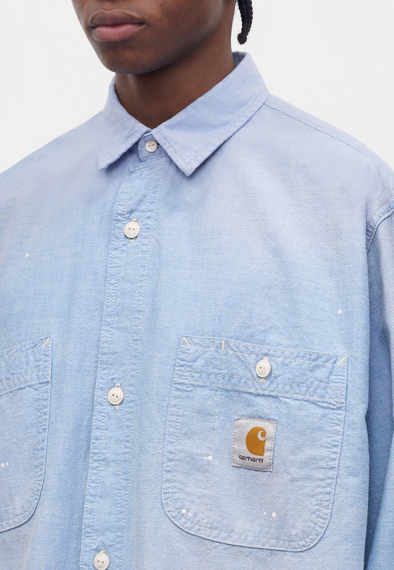 Persona che indossa una camicia azzurro chiaro con bottoni, tasche frontali e una toppa con il logo Carhartt sulla tasca sinistra, ritagliata al collo e alle spalle.