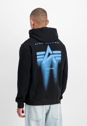 Homme aux cheveux courts et boucle d’oreille en anneau porte un sweat à capuche noir avec le logo bleu Alpha Industries et un jean clair, vu de dos sur fond uni.