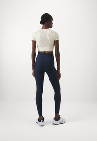 Svijetlo bež kratki rukav crop top s mrežastim detaljima, uparen s tamnoplavi visoki tajicama koje imaju logotip na nozi i bijelim tenisicama.