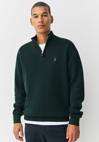 Joven vestido con un suéter verde oscuro de media cremallera con un pequeño emblema, camiseta blanca por debajo y pantalones oscuros, de pie frente a un fondo liso.