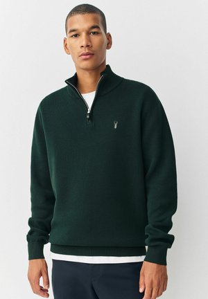 Young man wearing a dark green half-zip sweater with a small emblem, white undershirt, and dark pants, standing against a plain background.