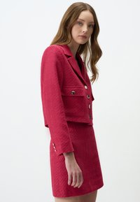 Veste courte rose vif en tissu texturé, avec deux poches avant et des boutons métalliques, assortie d'une jupe coordonnée. Fini lisse.
