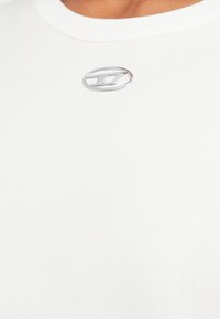 Balta kokvilnas T-krekls ar apaļu kakla izgriezumu un sudraba logotipu uz augšējās kreisās puses. Mīksta tekstūra ar gludu virsmu.