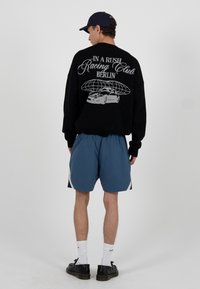 Czarny sweter z srebrnym nadrukiem graficznym przedstawiającym samochód i napis. Noszony z niebieskimi szortami, białymi skarpetkami i czarnymi butami.