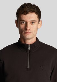 Mann mit kurzen lockigen braunen Haaren, der einen schwarzen Pullover mit Viertel-Reißverschluss und einem kleinen gestickten Logo auf der linken Brust trägt.