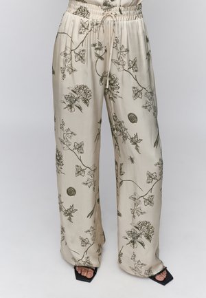 Pantaloni bej cu picior larg, cu talie elastică și șnur, decorați cu imprimeuri negre detaliate cu flori, viță de vie, melc și albine, asortați cu sandale negre.