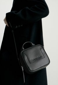 Černá kožená crossbody taška čtvercového tvaru, s vyraženým logem a zapínáním na zip. Obsahuje krátkou rukojeť a odnímatelný popruh.