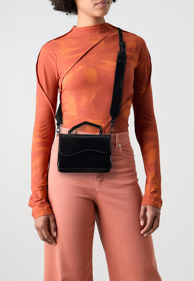 Sac à main noir en cuir embossé crocodile avec une poignée supérieure et une bandoulière réglable, présenté contre un haut orange à manches longues et un pantalon rose.
