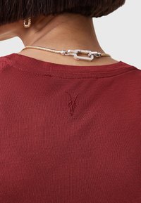 Czerwona, prążkowana bluzka z małym, haftowanym logo z tyłu przy szyi oraz metalowym łańcuszkiem z zapięciem. Gładka tekstura, dopasowany krój.