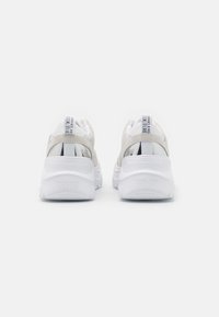 Bakvy av vita Versace Jeans Couture sneakers med beige mockadetaljer och märkta hälltag på enfärgad bakgrund.