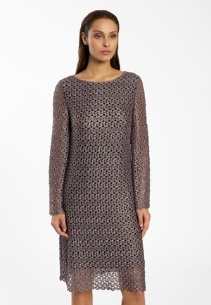 Vrouw die een paarse gehaakte kanten jurk draagt met lange mouwen en een lengte tot op de knie, met een ronde halslijn, staand tegen een effen witte achtergrond.