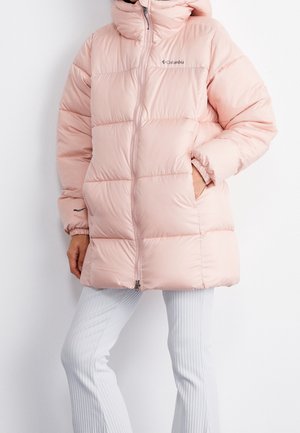 Rosa Steppjacke mit vorderem Reißverschluss und Logo auf der Brust, getragen über weißen Rippenhosen, Hände in den Taschen.