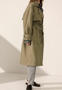 Olijfgroen trenchcoat met een ceintuur in de taille, knoopmanchetten en een kraag, gecombineerd met lichtgrijze jeans en donkere loafers.