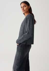 Femme aux longs cheveux foncés portant un haut long à manches grises texturé et un pantalon noir à jambes larges, posant de côté avec les mains dans les poches.