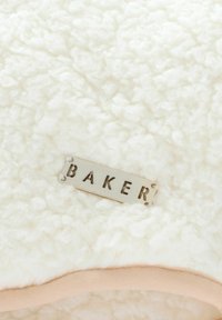 Tessuto bianco e soffice con una superficie texturizzata; un'etichetta metallica incisa con "BAKER" è attaccata a un bordo rosa pallido.