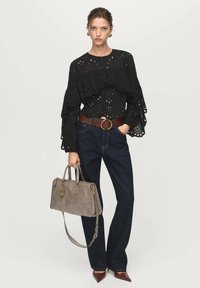 Blouse noire brodée avec des manches volantes, associée à un jean en denim foncé et un sac à main gris. Les accessoires comprennent une ceinture marron et des chaussures à bout pointu.