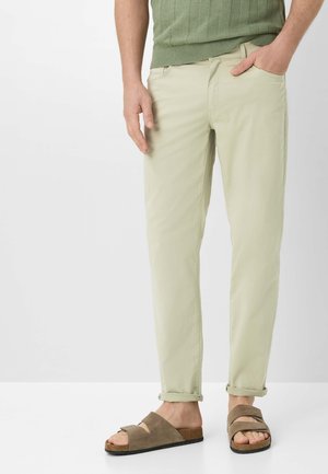 Mand iført lyse beige afslappede bukser med manchet og brune ruskind slip-on sandaler, stående mod en ensfarvet lys baggrund.