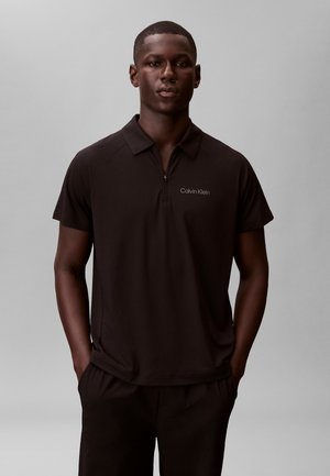 Mężczyzna w czarnej, krótkorękawowej koszulce polo Calvin Klein z kołnierzem na zamek, stojący na jednolitym szarym tle.