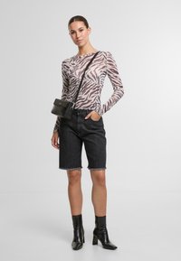 Mujer vestida con una blusa de manga larga de diseño de cebra transparente, pantalones cortos de mezclilla negros de cintura alta, botas negras hasta el tobillo y un bolso negro bandolera.