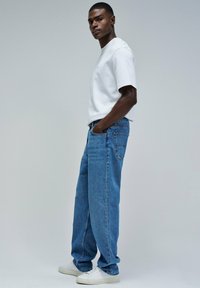 Camiseta blanca de cuello redondo, de corte relajado, combinada con jeans de denim azul claro que presentan una silueta holgada. Zapatillas blancas completan el atuendo.