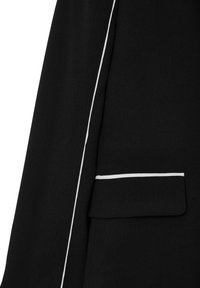 Tessuto nero con dettagli in piping bianco lungo le maniche e il bordo della tasca a tracolla di un indumento, probabilmente un blazer o una giacca.