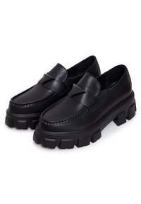 Chaussures mocassins noires, épaisses, avec des semelles texturées et des accents décoratifs triangulaires sur le devant supérieur.