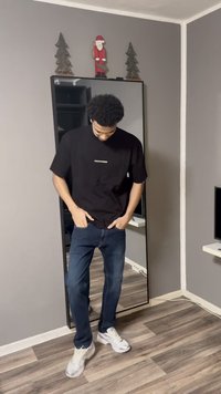 Crna majica kratkih rukava s logom "PEGADOG", plave traperice i bijele tenisice. Stoji ispred ogledala, gledajući dolje.