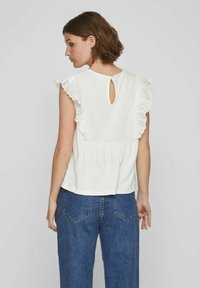 Top blanco con mangas de ganchillo con volantes y detalle de abertura en la parte posterior, combinado con jeans de mezclilla azul, que muestran un ajuste relajado y un estilo casual.