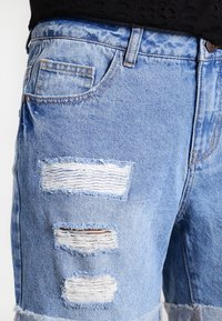 Shorts de mezclilla azul claro con parches desgastados, bordes deshilachados, cinco pasadores para cinturón y bolsillos en la parte delantera y trasera.