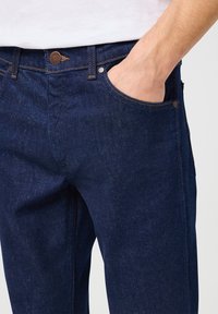 Donkerblauwe denim jeans met een rechte pasvorm, voorzien van contrasterende oranje stiksels en metalen accenten op de zakken en de tailleband.