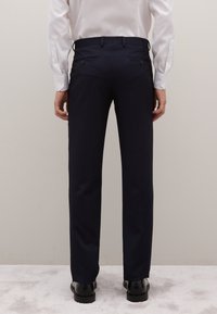 Pantalones de raya diplomática azul marino con un corte entallado, dos bolsillos traseros con cierre de botón y una textura suave. Combinados con una camisa blanca.