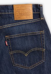 Mörkblå denimjeans med gul sömnad, en läderbaksida och en röd Levi's-bricka på bakfickan. Har klassisk fickdesign.