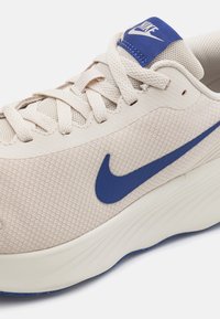 Chaussure de sport beige clair avec un dessus en maille, un swoosh bleu de Nike et des détails texturés. Elle possède un col rembourré et une semelle en mousse bien visible.