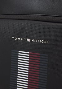 Zaino nero con texture che presenta il logo "TOMMY HILFIGER" e un design a strisce in rosso, bianco e blu su un lato.